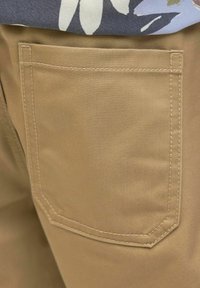 Pantalon en coton beige présentant une poche arrière avec une couture supérieure courbée, une fine texture verticale et des coutures contrastantes.