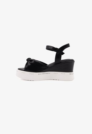 Czarne sandały na koturnie z węzełkowym przodem, syntetyczna cholewka, regulowany pasek wokół kostki oraz biała, teksturowana podeszwa.