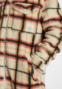Fleecejacke in Creme mit einem Karomuster, das rote, schwarze und orange Linien enthält. Mit Reißverschluss und einem Logo-Patch auf der Brust.