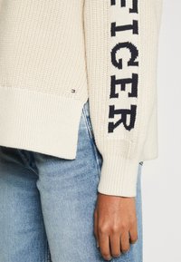 Pull en tricot crème avec le texte noir "TOMMY HILFIGER" sur la manche, porté avec un jean bleu clair ; main posée sur le côté.