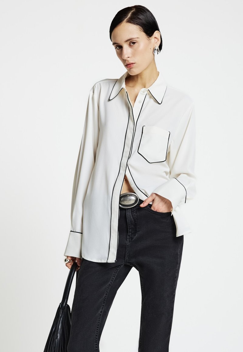 White long-sleeve shirt with black piping, featuring a front pocket and a classic collar, paired with high-waisted black jeans.