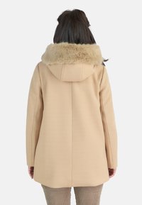 Abrigo beige con capucha, que presenta un suave collar de piel sintética. El material parece suave y estructurado, con un ajuste relajado y un corte recto.