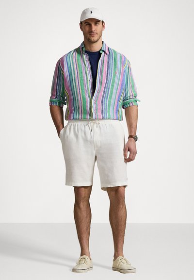 Chemise à rayures multicolores avec manches retroussées, assortie à un short blanc avec cordon de serrage et des baskets claires, le tout complété par une casquette blanche.