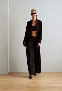 Black layered outfit featuring a long skirt and cropped top, made of lightweight fabric. Accompanied by dark sunglasses and a clutch.