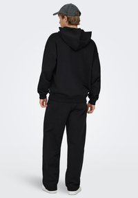 Schwarzer Pullover mit Kapuze, großzügiger Kapuze, gerippten Bündchen und lockerem Schnitt. Kombiniert mit passenden schwarzen Sweatpants und weißen Sneakern.