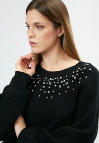 Schwarzer Pullover mit gerippter Textur, ausgestattet mit einem mit Juwelen besetzten Ausschnitt in verschiedenen Größen und Formen. Lange Ärmel und ein Rundhalsausschnitt-Design.