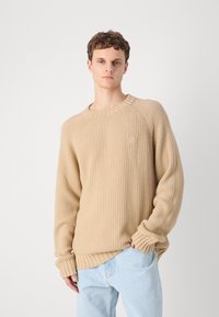 Beige ribbed knit sweater with a crew neck, raglan sleeves, and a small logo detail on the chest. Paired with light blue jeans.