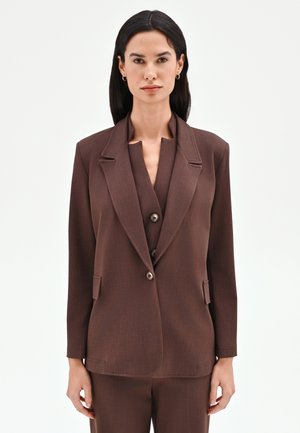 Donna con capelli lisci e scuri che indossa un blazer marrone su misura e pantaloni abbinati, in piedi davanti a uno sfondo chiaro e uniforme.