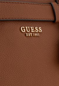 Brown textured synthetic leather handbag featuring a gold "GUESS EST 1981" logo and black stitching accents.