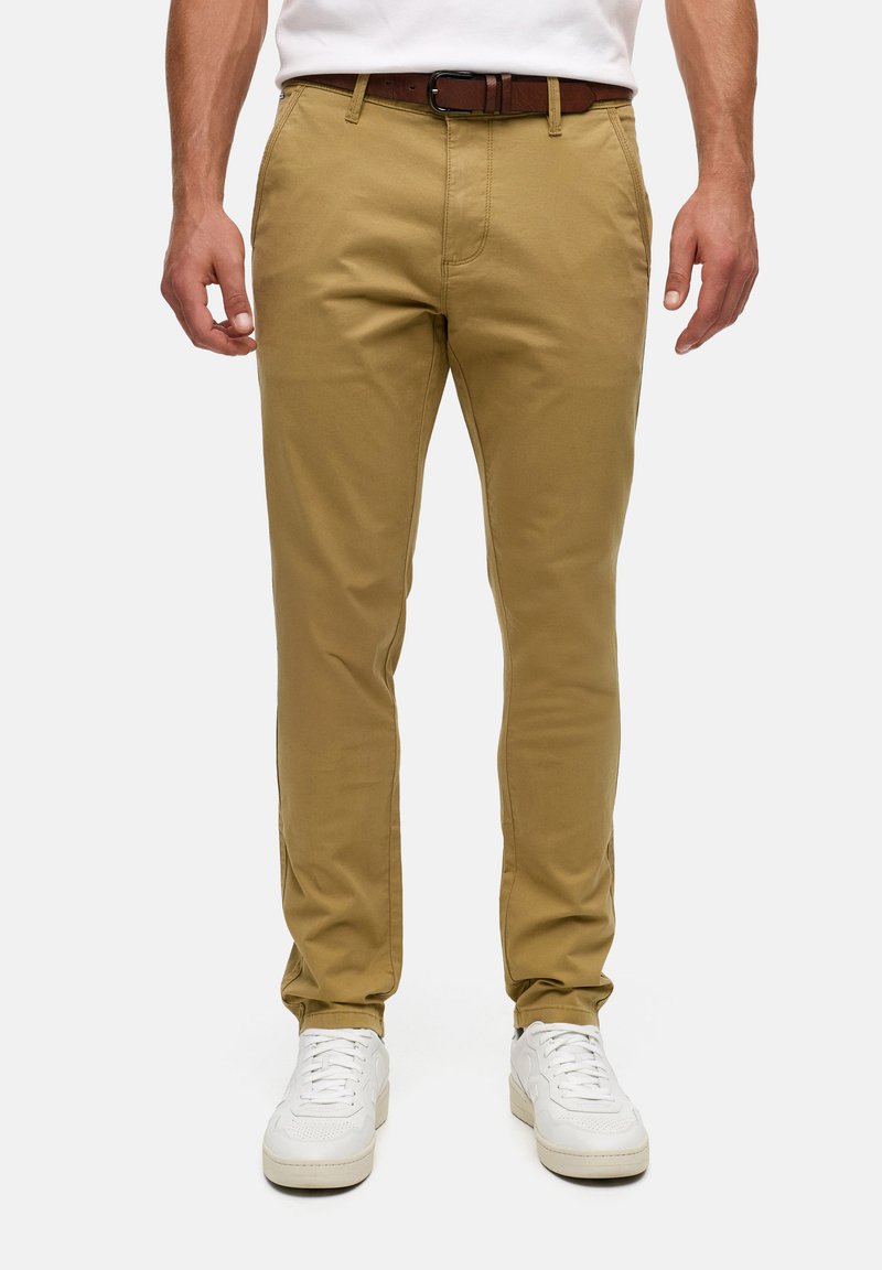 Homme portant un pantalon kaki ajusté avec une ceinture marron et des baskets blanches, debout devant un fond clair uni.