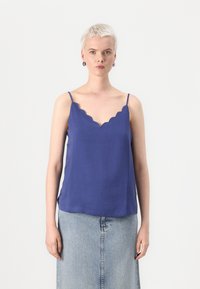 Top in raso viola con bordi scallop, realizzato in tessuto leggero, caratterizzato da spalline sottili e una vestibilità ampia, abbinato a una gonna di denim chiaro.