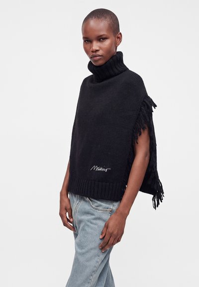 Personne portant un poncho noir à franges avec un col roulé et un logo blanc, ainsi qu'un jean bleu clair, se tenant devant un fond uni clair.