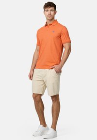 Polo shirt orange à manches courtes avec une texture subtile et un petit logo, associé à un short beige et des baskets blanches.
