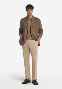 Brun kavaj över en beige polo, kombinerad med beiga byxor och svarta skor. Släta texturer, åtsittande design, en liten fickdetalj på kavajen.