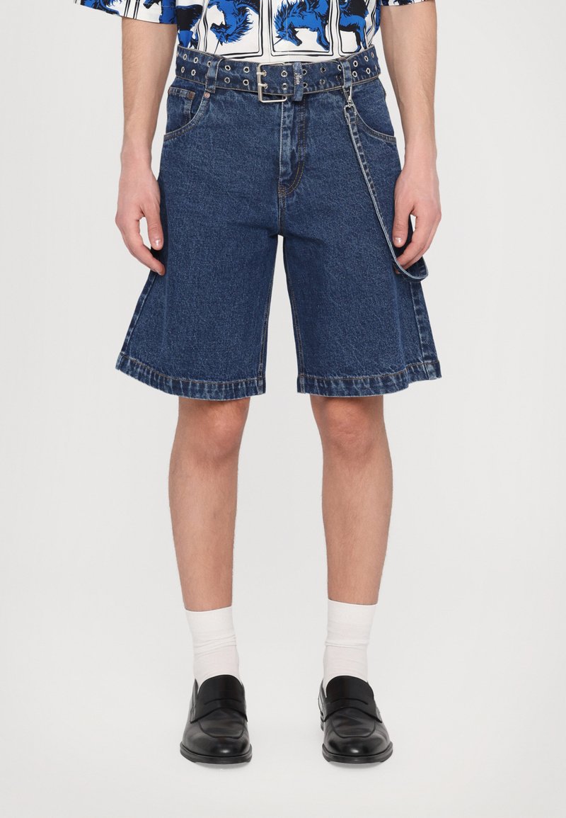 Șorturi din denim cu o croială lejeră, de culoare albastră, cu picioare largi și margine brută. Prezentând un centură cu ținte și un accesoriu cu lanț. Purtați cu pantofi negri și șosete albe.