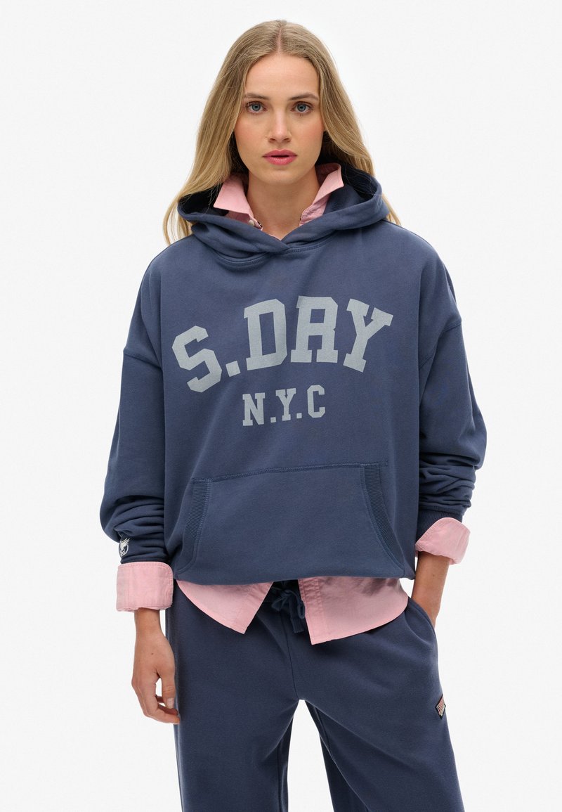 Sudadera azul marino con texto gris "S.DAY N.Y.C.", bolsillo tipo canguro y puños acanalados, superpuesta sobre una camisa de collar rosa claro.