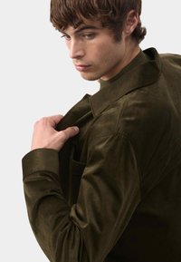 Oliwkowa koszula z korduru z kieszonką na piersi, z kołnierzykiem i zapięciem na guziki przy mankietach. Materiał ma teksturowany, prążkowany wygląd.