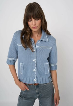 Kobieta ubrana w jasnoniebieski, krótki sweter-kardigan z białymi przeszyciami i białymi guzikami, zestawiony z niebieskimi dżinsami i brązowym paskiem.