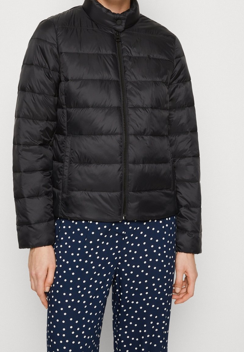 Giacca a vento nera con zip frontale, dotata di colletto alto. Il tessuto ha una texture liscia con cuciture orizzontali. Indossata con pantaloni a pois.