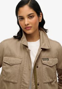 Camelkleurig katoenen jack met een kraag, twee borstzakken en een rits aan de voorkant. Voorzien van lichte stiksels en een klein logopatch.