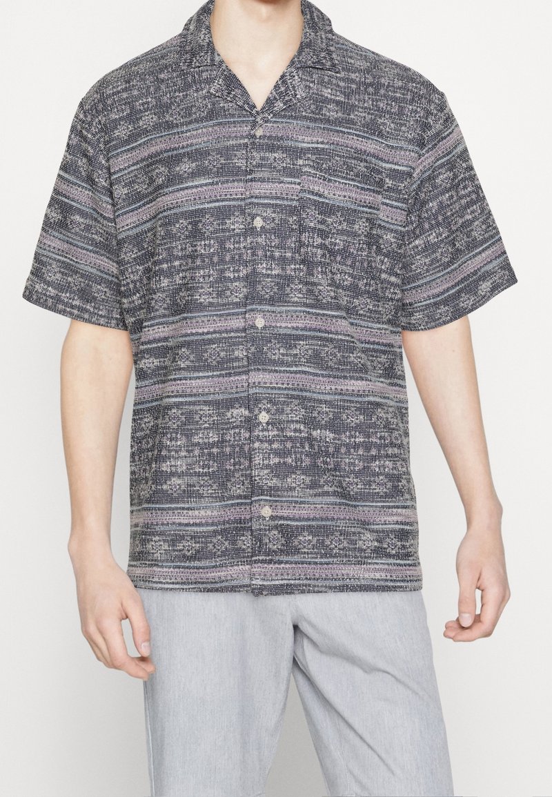 Homme portant une chemise à manches courtes boutonnée avec des rayures horizontales à motifs noirs, blancs et violets, associée à un pantalon gris clair.