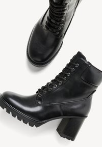 Bottines en cuir noir avec fermeture à lacets à l'avant, talons blocs épais et semelles robustes sur fond blanc.