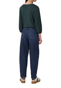 Donkergroene top met textuurmouwen en losvallende blauwe jeans. Model draagt wit en zwarte sneakers. Gezicht vanaf de achterkant.