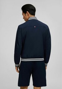 Marine bomberjack in een gladde stof met witte gestreepte ribgebreide mouwen en zoom. Bevat een klein rood, wit en blauw logo op de achterkant van de hals.