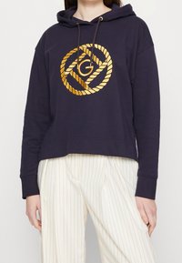 Sudadera con capucha de color navy con un logotipo de diseño de cuerda dorada en el frente. Tejido suave, corte cropped y puños acanalados. Combinada con pantalones ligeros a rayas.
