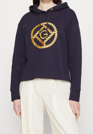 Sudadera con capucha de color navy con un logotipo de diseño de cuerda dorada en el frente. Tejido suave, corte cropped y puños acanalados. Combinada con pantalones ligeros a rayas.