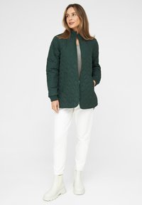 Gestepptes, smaragdgrünes Jacket mit Stehkragen, zwei vorderen Taschen und gerippten Bündchen, getragen über einem hellgrauen Oberteil und weißen Hosen.