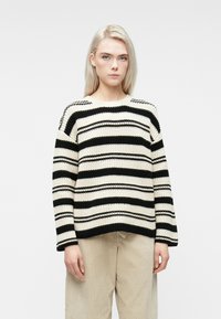 Schwarz-weiß gestreifter Pullover mit breitem Rundhalsausschnitt, grobgestrickter Textur und überschnittenen Schultern. Kombiniert mit beigen Cordhosen.