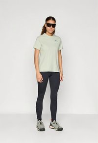 Lichtgroen T-shirt met een klein blauw logo, zwarte legging en grijze sneakers met roze accenten; model draagt een zonnebril en staat rechtop.