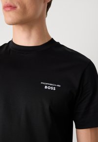 Melns kokvilnas T-krekls ar apaļu kaklu, kurā kreisajā krūtīs ir balta izšūta "Porsche" logo un "BOSS" teksts.