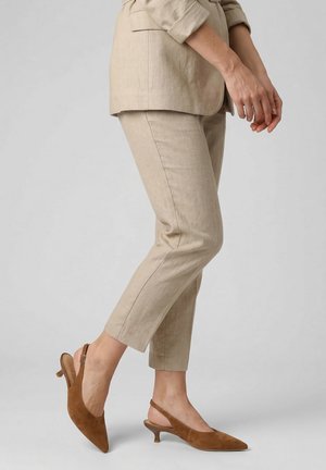 Vrouw draagt beige cropped broek en bijpassende blazer, poserend in bruine suède slingback kitten heel schoenen tegen een lichtgrijze achtergrond.