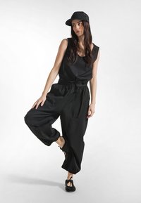 Schwarzes ärmelloses Oberteil kombiniert mit lockeren, schwarzen, mit Gürtel versehenen Hosen mit Taschen und gerafften Beinabschlüssen; schwarze Mütze und schwarze Slip-On-Schuhe.