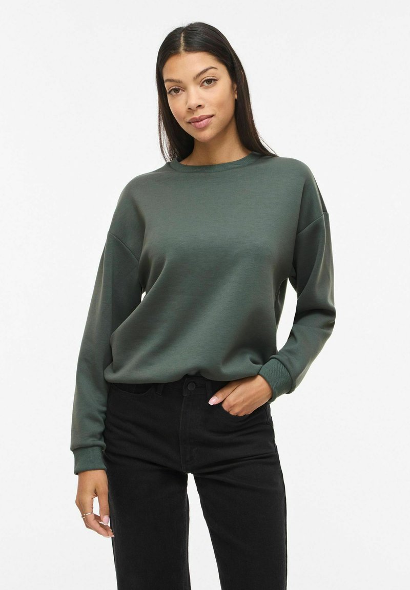 Femme aux longs cheveux foncés portant un sweat-shirt vert foncé à col rond et un pantalon noir, debout avec une main dans la poche sur fond blanc.