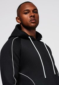 Selbstbewusster Mann mit kurzen Haaren, der einen schwarzen Hoodie mit weißen Nähten und Kordeln vor einem einfarbigen hellen Hintergrund trägt.