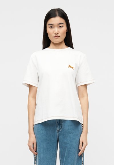 Jeune femme aux longs cheveux bruns portant un t-shirt blanc avec un petit chien brodé et un jean bleu avec des ornements latéraux.