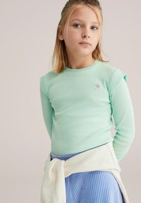 Mentaszínű bordás hosszú ujjú felső vállrészletes és apró virágos hímzéssel, világoskék bordás szoknyával és deréknál megkötött krém színű pulóverrel párosítva.