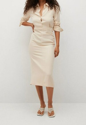 Woman wearing a beige long-sleeve buttoned dress and white open-toe heeled sandals standing against a plain light background.