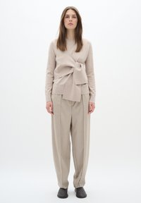 Beige Wollmischung Pullover mit einem Knoten-Detail vorne, kombiniert mit hellbeigen hochtaillierten Hosen und dunklen Schuhen vor einem weißen Hintergrund.