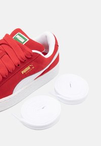 Baskets en suède rouge avec semelle en caoutchouc blanc et rayure. Elles possèdent de grosses lacets rouges et une étiquette logo verte. Comprend deux lacets blancs roulés.