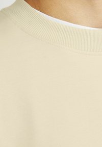 Sudadera de algodón beige con un escote acanalado y textura suave; diseño sin costuras que presenta un corte simple y limpio en el cuello.