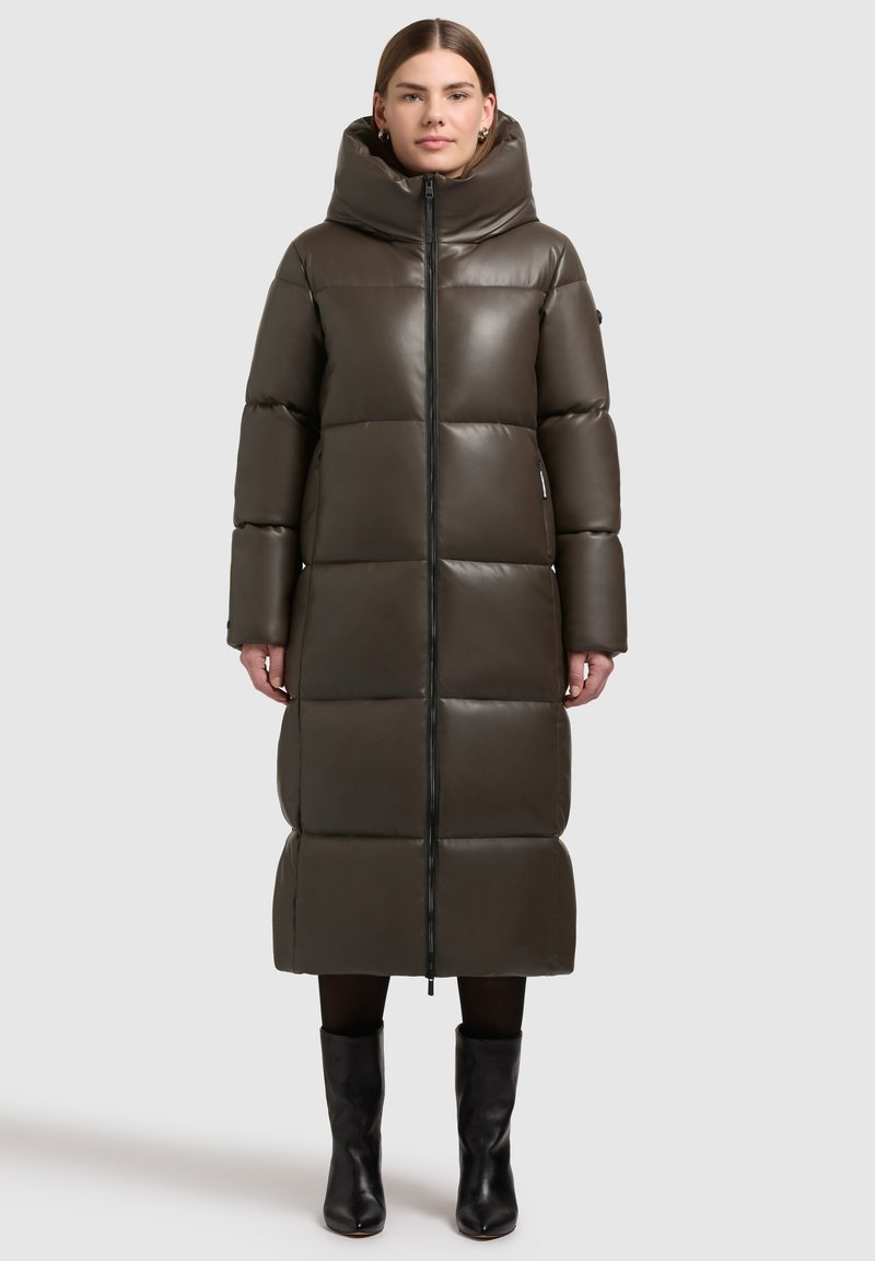 Geacă puffer lungă, de culoare verde oliva, cu guler înalt, design matlasat, fermoar frontal și mâneci lungi, asociată cu cizme negre până la genunchi.