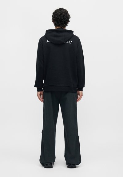 Άνδρας με γκρίζα μαλλιά που φοράει μαύρο φούτερ με κουκούλα με μερική λευκή επιγραφή στο πάνω μέρος της πλάτης, μαύρο παντελόνι με φαρδιά μπατζάκια και μαύρα παπούτσια, στραμμένος προς τα πίσω.