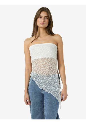 Femme portant un top en dentelle blanche sans bretelles avec un ourlet asymétrique, associé à un jean bleu, debout devant un fond blanc uni.