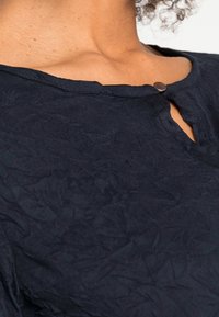 Granatowa bluzka z teksturowanej tkaniny, z małym wycięciem w kształcie łezki przy dekolcie i okrągłym metalowym guzikiem jako akcentem.