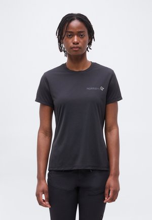 Νεαρός με κοντά ντραντλοκς, φορώντας εφαρμοστό μαύρο T-shirt και μαύρο παντελόνι, στέκεται μπροστά σε απλό φωτεινό φόντο.