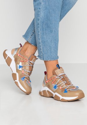 Osoba nosząca beżowo-białe masywne trampki z niebiesko-pomarańczowymi sznurówkami, w zestawie z jasnoniebieskimi, skróconymi dżinsami na neutralnym tle.